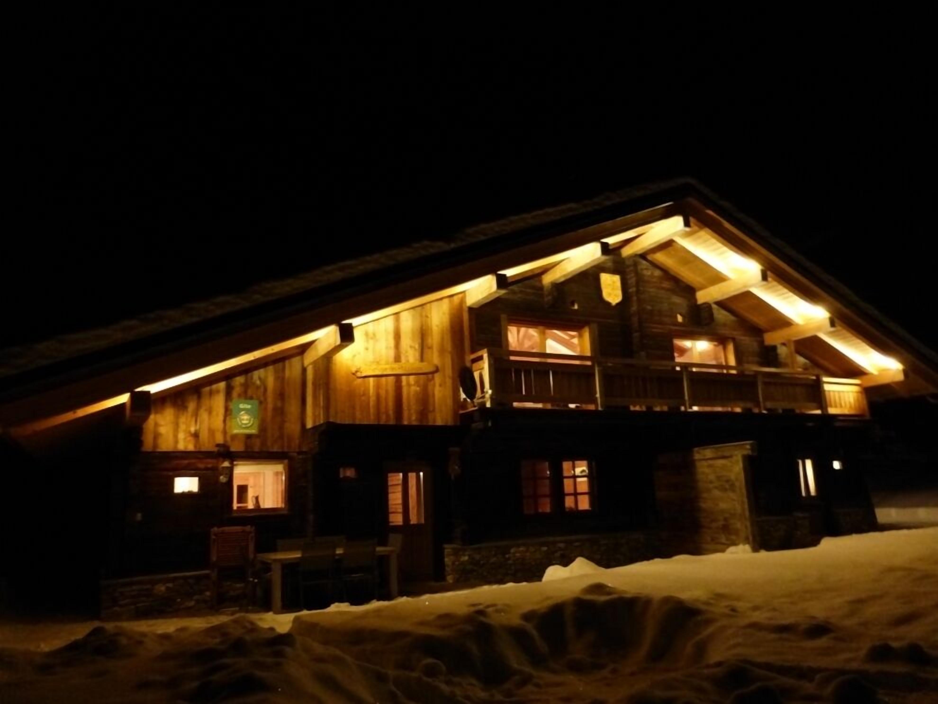 Una cabaña de troncos con luz cálida en su interior, fachada de madera y suelo cubierto de nieve.