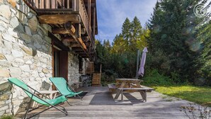 Une terrasse en bois avec deux fauteuils de jardin verts et une table de pique-nique, entourée de murs en pierre et d’une zone boisée.