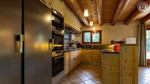 Une cuisine compacte avec des meubles en bois, un réfrigérateur en acier inoxydable et un four encastré. Des luminaires suspendus et une fenêtre laissent entrer la lumière naturelle.