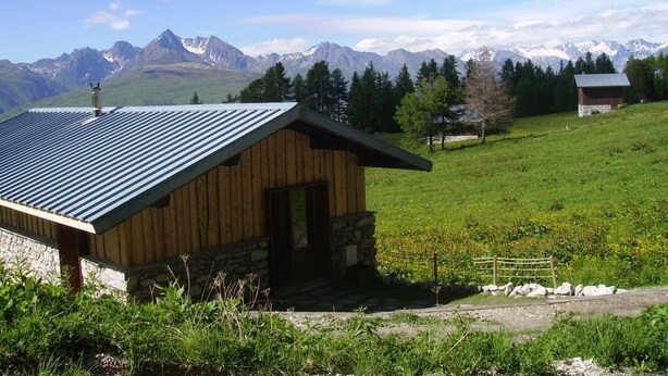 Un edificio de madera rústico con techo de metal inclinado, ubicado en una ladera cubierta de hierba y con montañas nevadas al fondo.