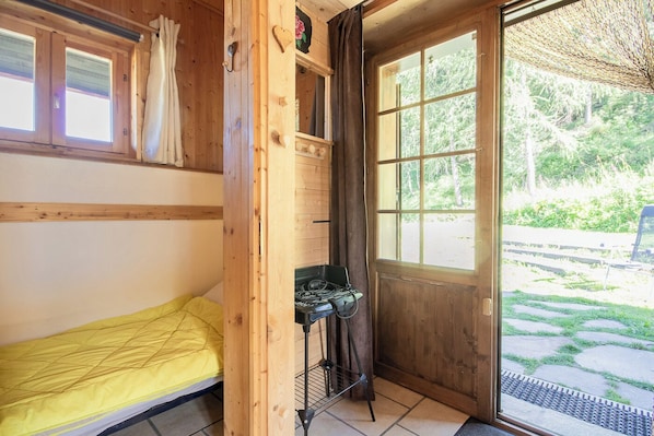 Eine Holzhütte mit einem Einzelbett, einer kleinen Küchenzeile und einem großen Fenster mit Blick ins Freie.