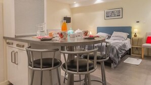Habitación de hotel compacta con kitchenette, zona de comedor con taburetes de bar, cama con cabecera azul y mesita de noche con lámpara.