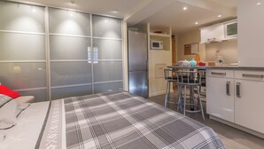 Un dormitorio moderno con cama, armario corredero, kitchenette con microondas y comedor con mesa y sillas.