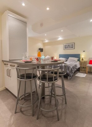 A compact hotel room with a kitchenette, a dining area with bar stools, a bed with a blue headboard, and a bedside table with a lamp.