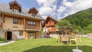 A two-story chalet with a stone entrance, wooden balconies, and a grassy yard with outdoor seating. - LA Ferme DES Moulins (VALLOIRE)