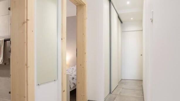 Een smalle gang met houten deuren, een bed met een zwart-wit geruite sprei en een betegelde vloer.