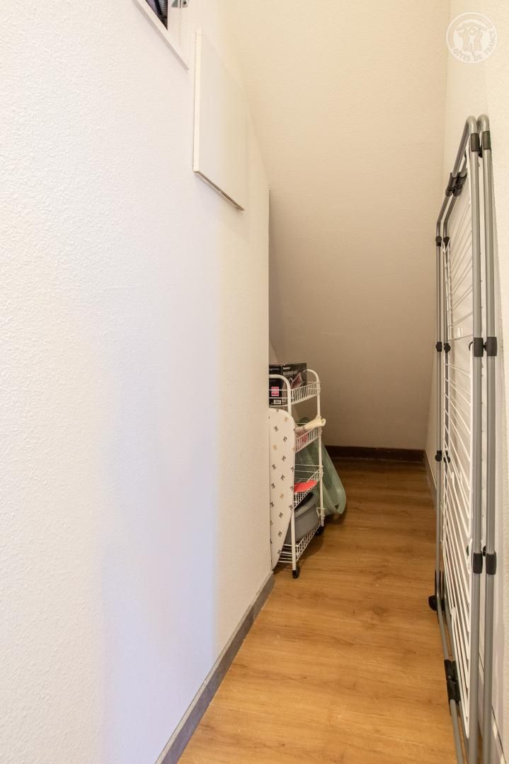 A narrow hallway with a wooden floor, a white wall, and a metal rack with storage bins.