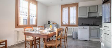 A modern kitchen with white cabinets, a built-in oven, and a dishwasher. There is a dining area with a wooden table and chairs, and a window with curtains.