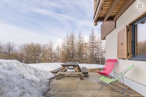 Eine Holzterrasse mit Picknicktisch und zwei Stühlen, eine verschneite Landschaft und ein Gebäude mit Fenster und Holzläden.