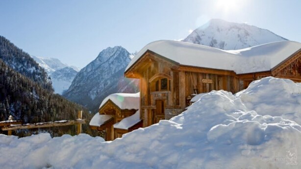 A wooden lodge with a snow-covered roof, surrounded by snow and mountains.