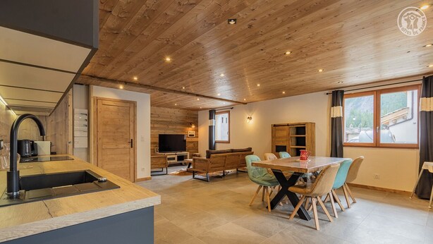 A modern kitchen with a built-in oven and sink, a dining area with a wooden table and chairs, and a living space with a TV and seating.