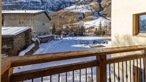 A snow-covered courtyard with a wooden balcony, stone buildings, and a mountain backdrop.