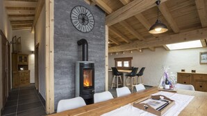 Un interior moderno con una pared de piedra, una estufa de leña, una zona de comedor con una mesa larga y una cocina con taburetes de bar.