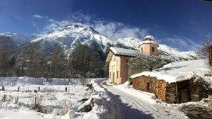 Een besneeerd berglandschap met een klein gebouw met een klokkentoren, omringd door bomen en een besneeuwde pad.