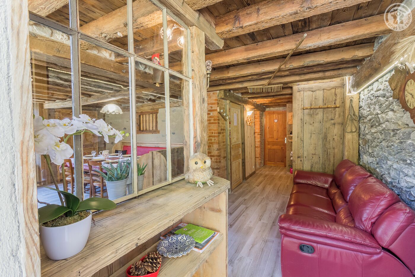 Un intérieur rustique avec un plafond en bois, un canapé en cuir rouge, un miroir reflétant un coin repas et un mur en pierre.