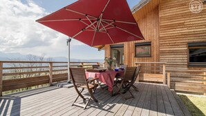 Een houten terras met een tafel, stoelen en een rode parasol, met uitzicht op bergen.