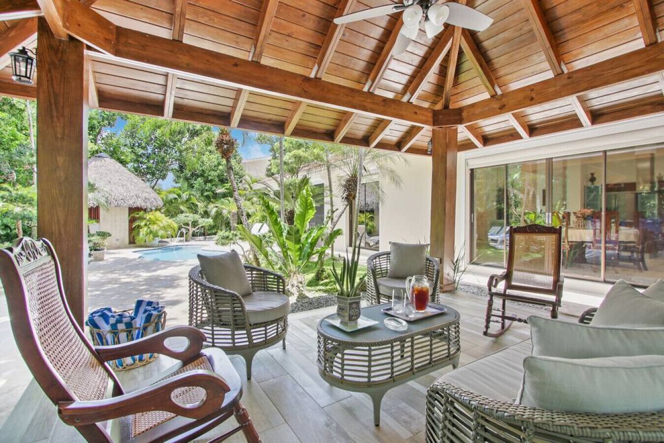 Un espacio al aire libre techado con mobiliario de mimbre, una mesa con jarras y vistas a una piscina y jardín.