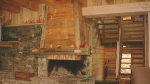 Interior rústico de una cabaña de troncos con chimenea de piedra, vigas de madera y una zona de estar acogedora.