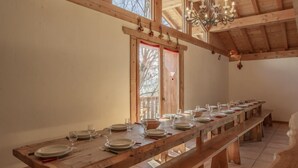 Una larga mesa de comedor de madera dispuesta con platos, vasos y cubiertos, en una habitación con vigas de madera y un candelabro.