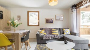 Una sala de estar moderna con un sofá gris, cojines amarillos y grises, una mesa de centro blanca y un comedor con una mesa y sillas de madera.