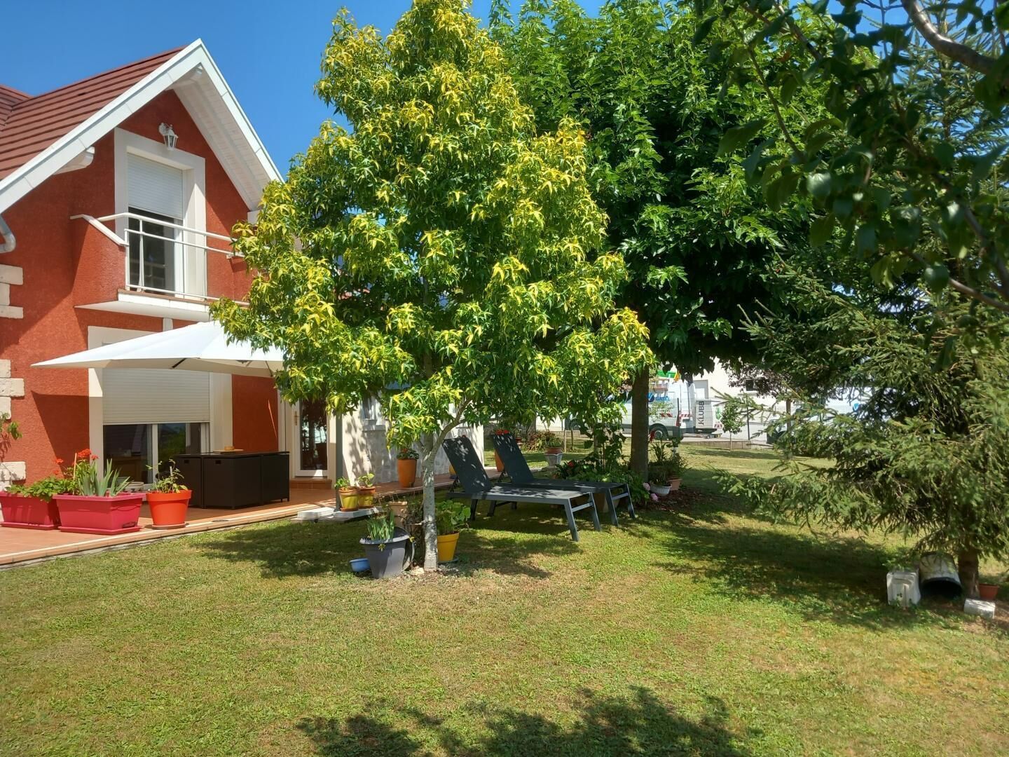A house with a red exterior, a white roof, and a covered patio area. There is a tree with yellow leaves in the foreground, and a lawn with a few potted plants and a chair.