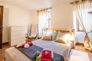 Quarto bem iluminado com cama de madeira, uma cadeira, mesa de cabeceira e janela com cortinas.