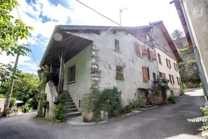 Um edifício de pedra tradicional com telhado inclinado, uma escadaria que leva à entrada e um pequeno jardim com plantas em vasos.