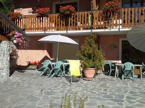 Une terrasse extérieure avec un sol en pierre, des chaises vertes, une table avec un parasol blanc et un arbre en pot.