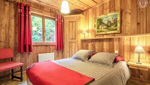 A wooden room with a bed, a chair, a painting, and a window with curtains.