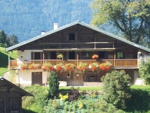 Een houten chalet met een balkon, omgeven door weelderige begroeiing en bergen.
