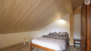 Une chambre avec un plafond en pente, un plancher en bois, un lit avec une literie à motifs, une chaise et une armoire en bois.