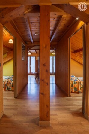 A wooden hallway with a high ceiling and a central wooden column. The hallway leads to a room with a bed and a window. - LA Grange Villard - Albiez Montrond (ALBIEZ MONTROND)