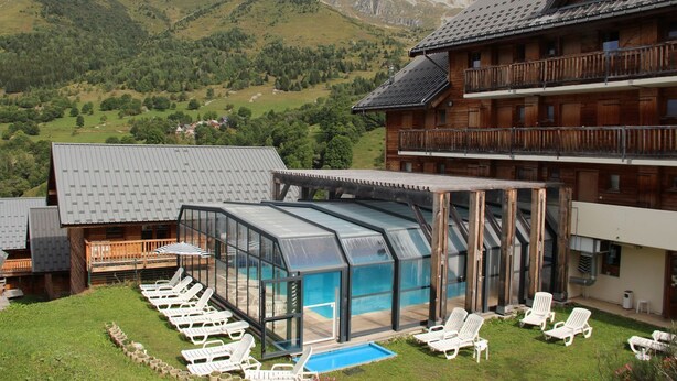 Una cabaña con piscina cubierta de cristal, sillones blancos y montañas al fondo.