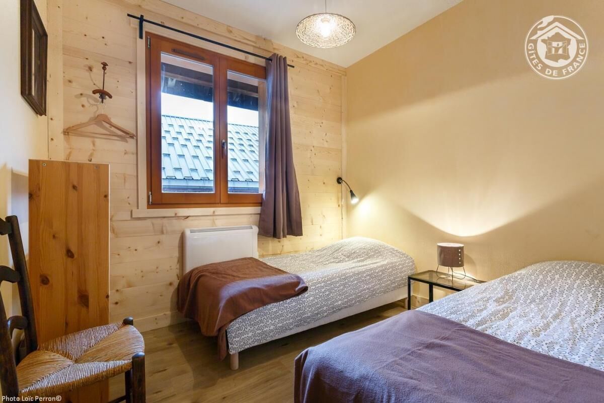 Une petite pièce chaleureuse aux murs en bois et au plafond en pente, comprenant deux lits simples, une chaise, une table et un porte-manteau mural.