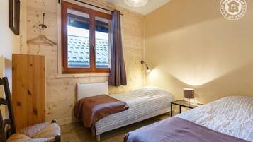 Une petite pièce chaleureuse aux murs en bois et au plafond en pente, comprenant deux lits simples, une chaise, une table et un porte-manteau mural.