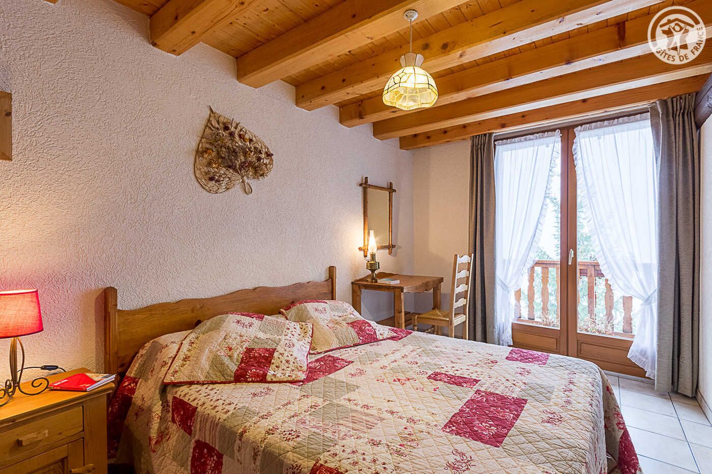 Un dormitorio con techo de madera, cama con colcha floral, escritorio con lámpara y ventana con cortinas.