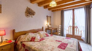 A bedroom with a wooden ceiling, a bed with a floral quilt, a desk with a lamp, and a window with curtains. - LES Diablotins - St-francois-longchamp (SAINT FRANCOIS LONGCHAMP)