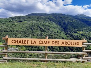 Ein Holzschild mit der Aufschrift 'CHALET LA CIME DES AROLES' vor einer gebirgigen Landschaft.