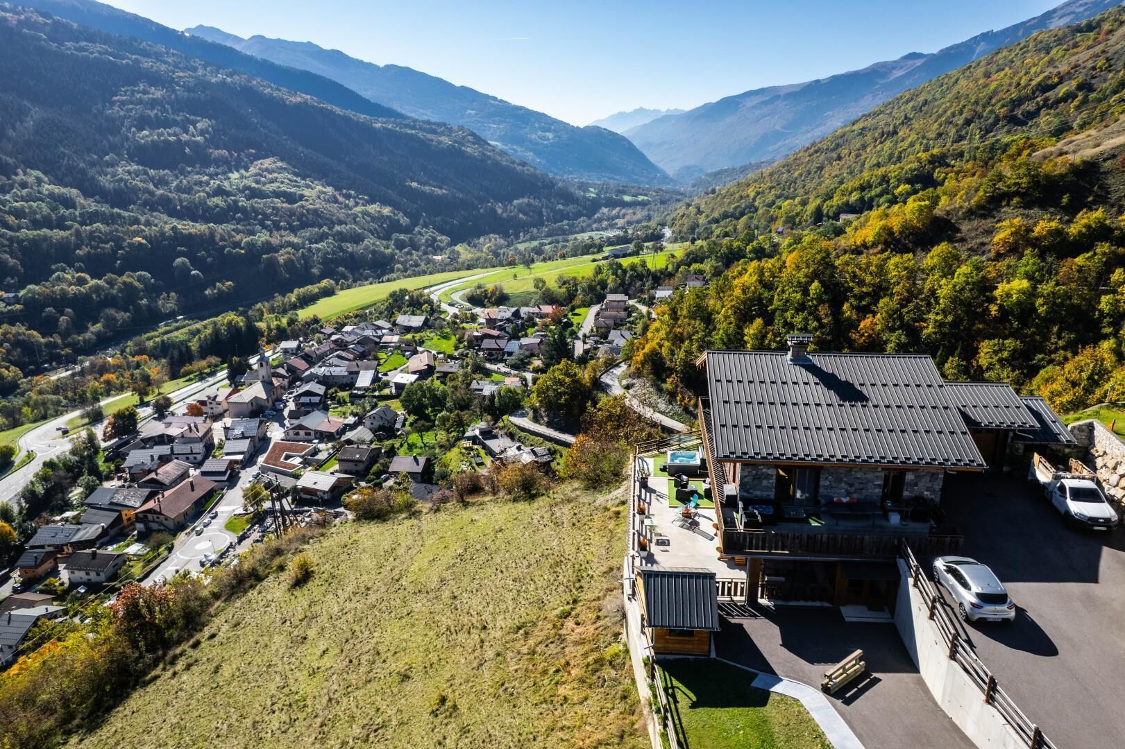 A mountain village with a central road, houses, and a large building with a sloped roof and outdoor seating area.