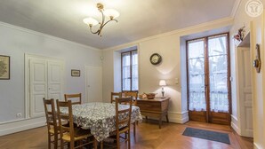 A dining area with a table and chairs, a chandelier, a clock, and a window with lace curtains.