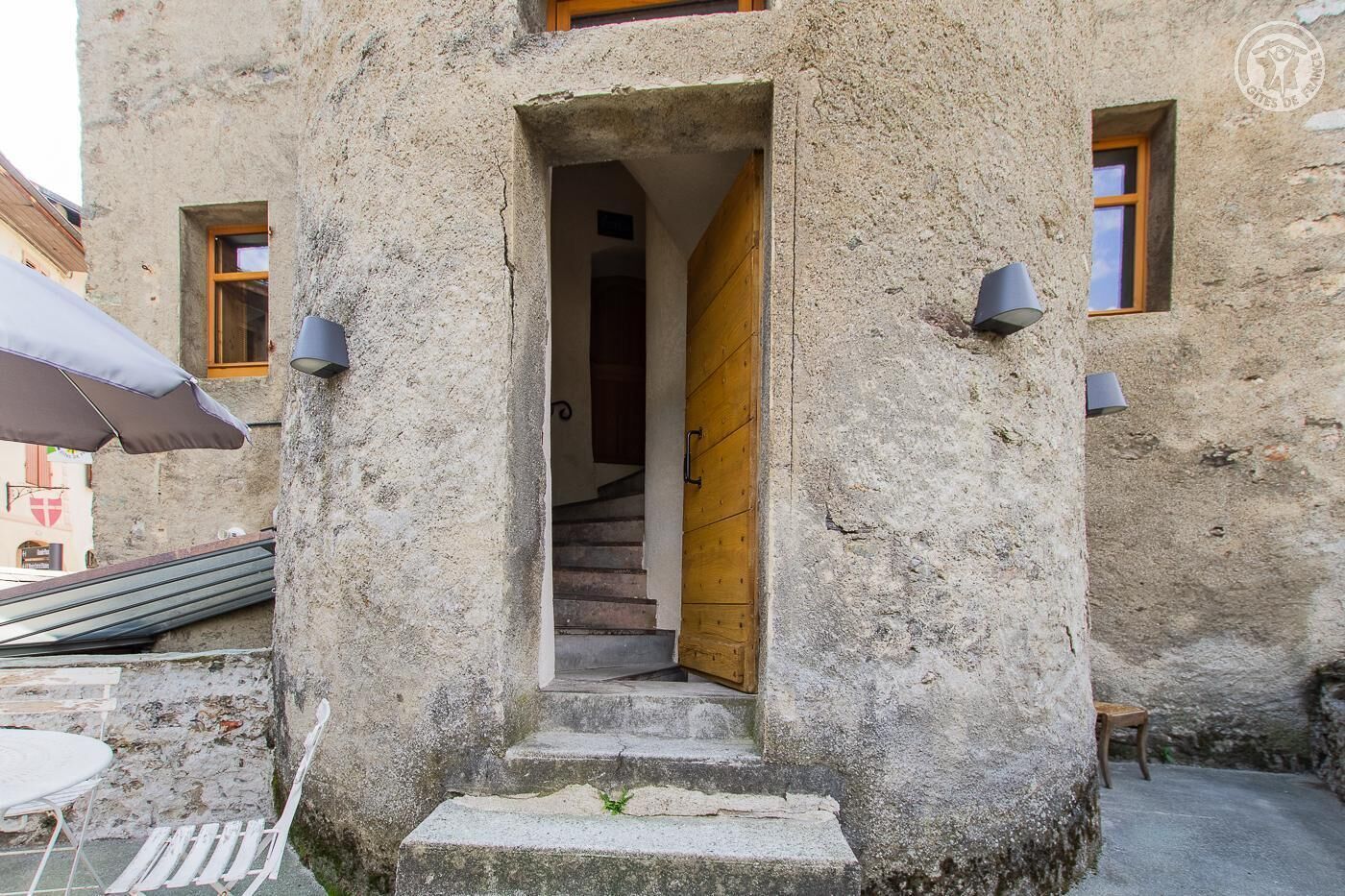 A stone building with a wooden door, a staircase leading inside, and two wall-mounted light fixtures.