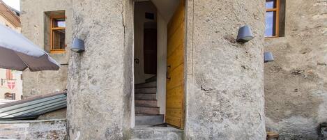 A stone building with a wooden door, a staircase leading inside, and two wall-mounted light fixtures.