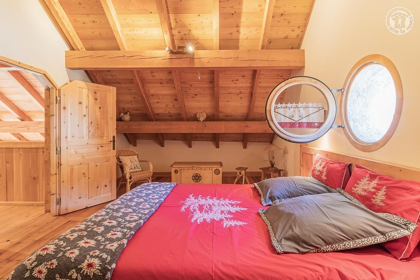 Un dormitorio con techo de madera, una cama con ropa de cama rojo y gris, un espejo redondo y una ventana con vistas.