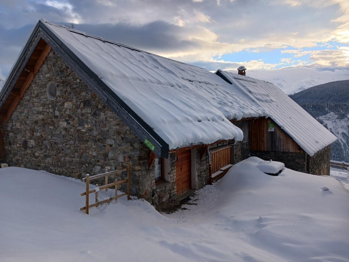 Un bâtiment en pierre avec un toit en pente recouvert de neige, des portes en bois et une clôture en bois.