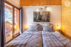 A cozy bedroom with two beds, a large window with curtains, a painting above the beds, and wooden walls.
