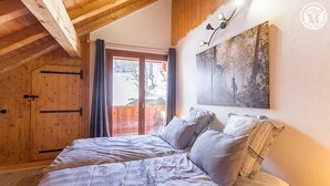 A bedroom with a wooden ceiling, a bed with patterned bedding, a painting on the wall, and a view of a snowy landscape through a door.