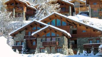 Un chalet à étages doté de balcons en bois et de murs en pierre, entouré d’arbres couverts de neige.