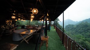 An outdoor seating area with a view of a forested hillside, wooden furniture, and hanging light fixtures. - Mudras Grove Resort (Nainital)