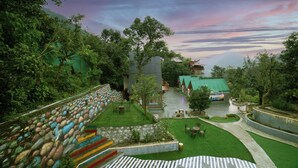 A landscaped garden with a stone wall, a grassy area, and a pathway leading to a building with a green roof. - Mudras Grove Resort (Nainital)