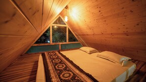 A cozy attic bedroom with a wooden sloped ceiling, a large window, and a bed with a patterned rug. - Mudras Grove Resort (Nainital)